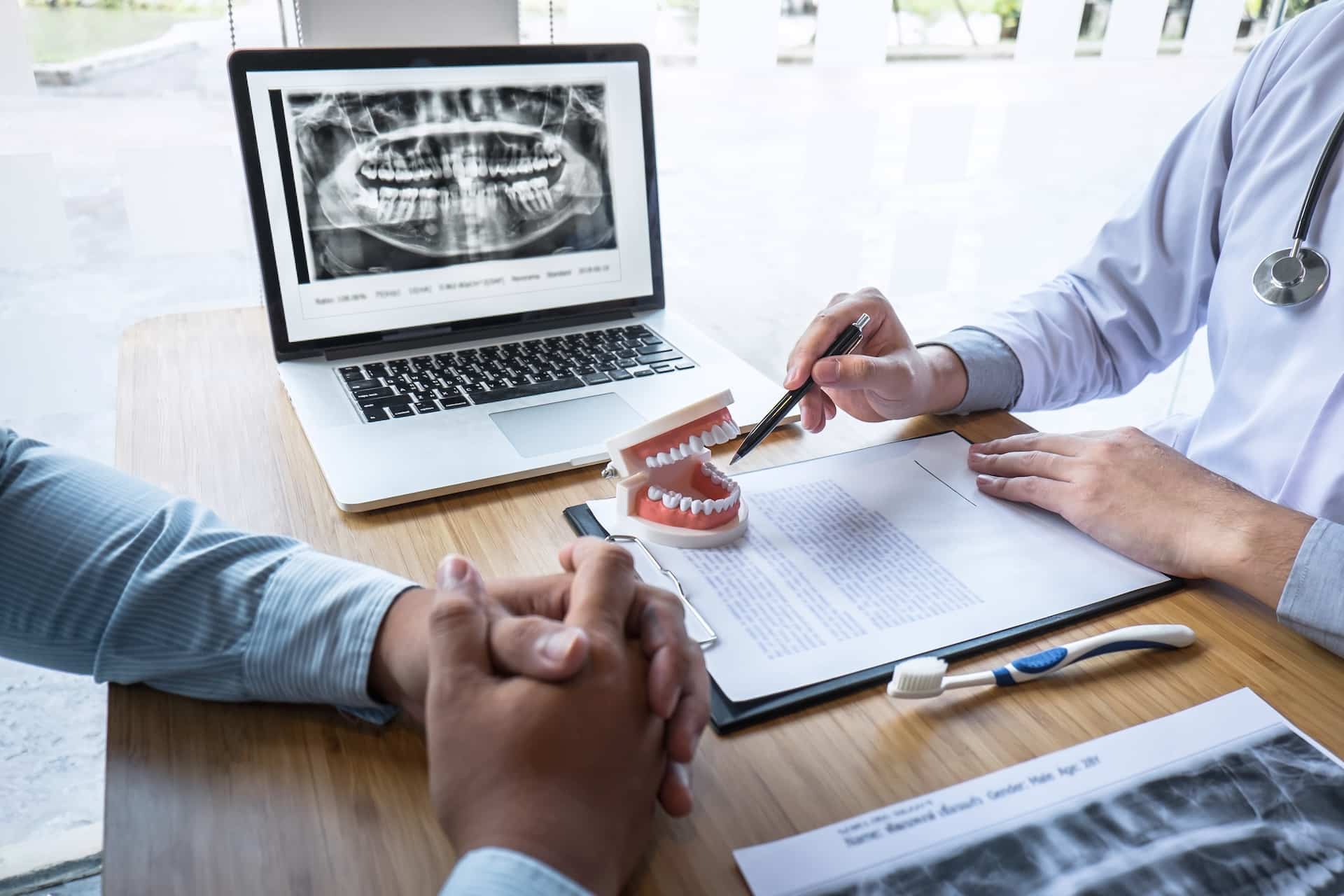 Two people sit at a table discussing dental X-rays displayed on a laptop screen. One person uses a dental model and pen to explain, while mentioning details about dental insurance. A toothbrush and documents are visible on the table.