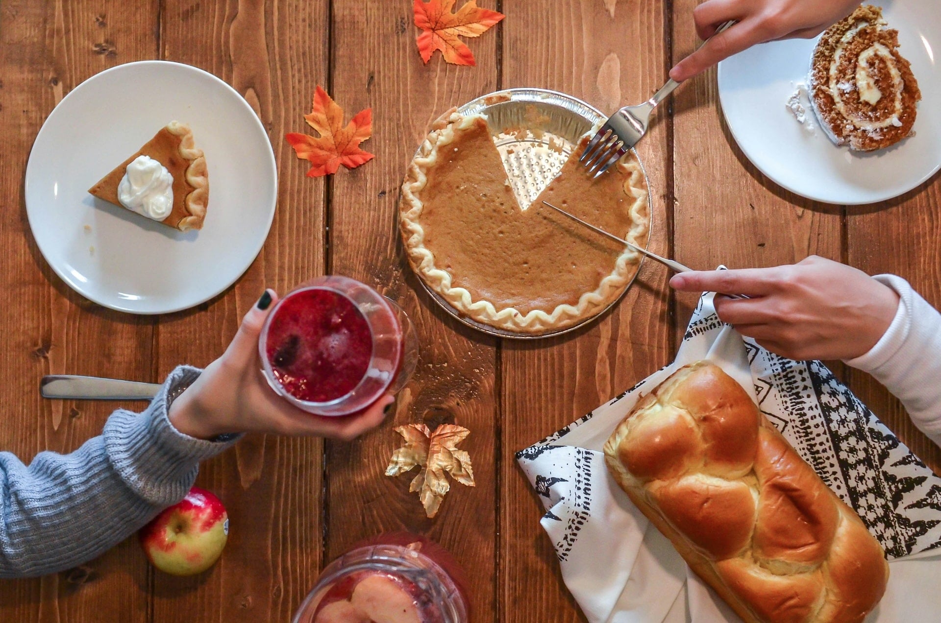 A top-down view of a table displays an inviting pumpkin pie, a slice topped with whipped cream, braided bread, an apple, a beverage, and autumn decorations.